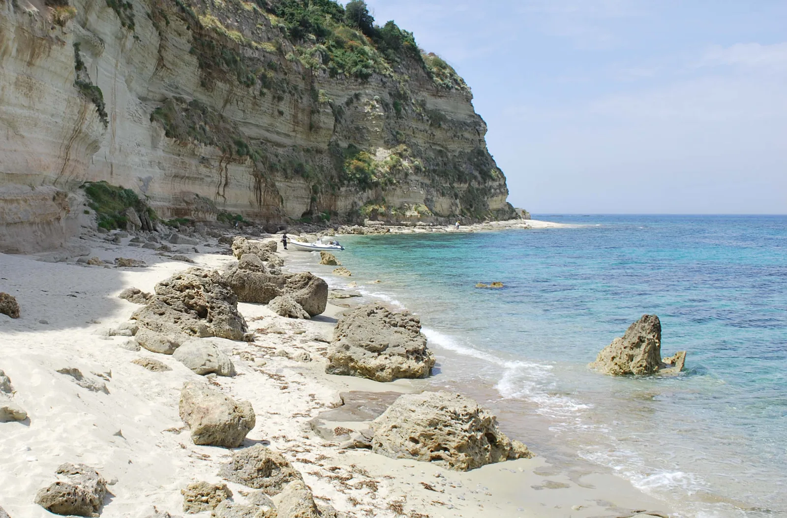 Spiaggia selvaggia con rocce, sabbia chiara e imponente falesia sul mare azzurro