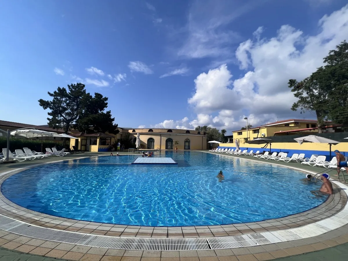Piscina circolare con bagnanti, lettini bianchi e cielo azzurro