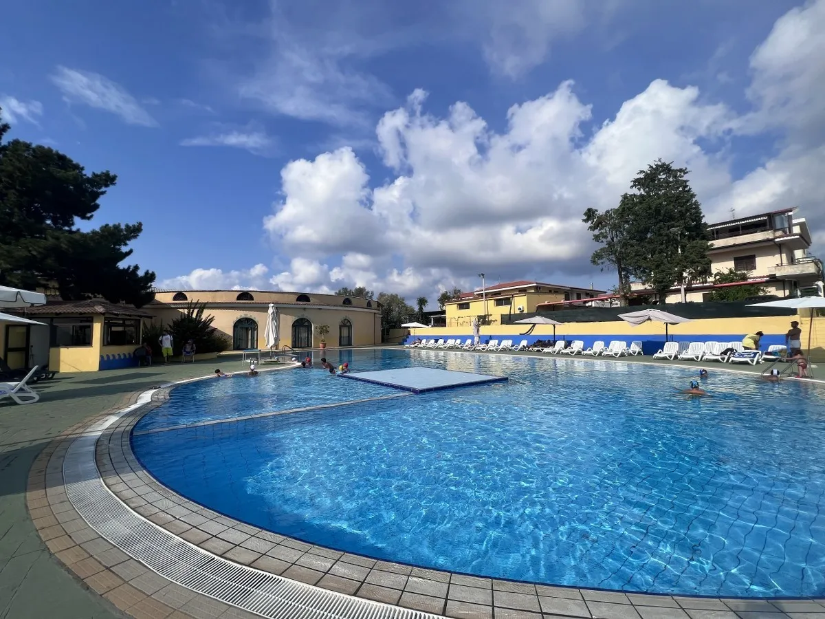 Piscina con piattaforma centrale, lettini e bagnanti sotto cielo nuvoloso