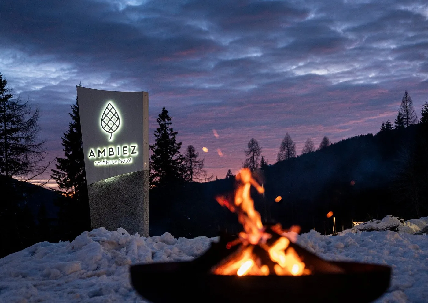 Insegna illuminata dell'hotel Ambiez con falò acceso nella neve al tramonto