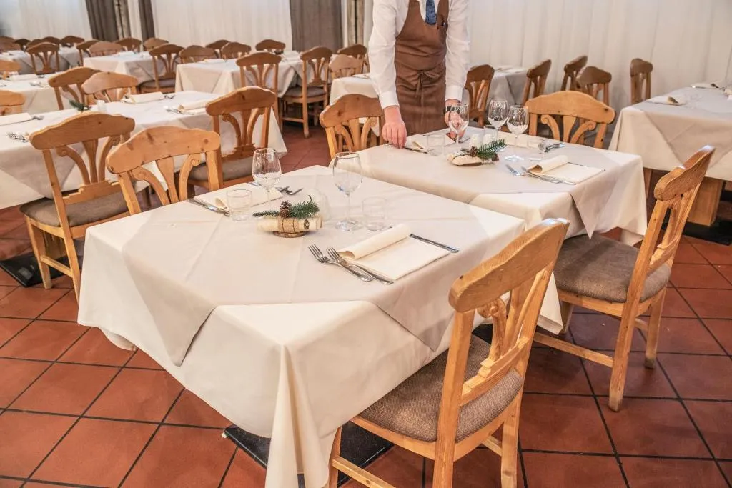 Sala ristorante con tavoli vestiti di tovaglie bianche, sedie in legno e cameriere che prepara la mise en place