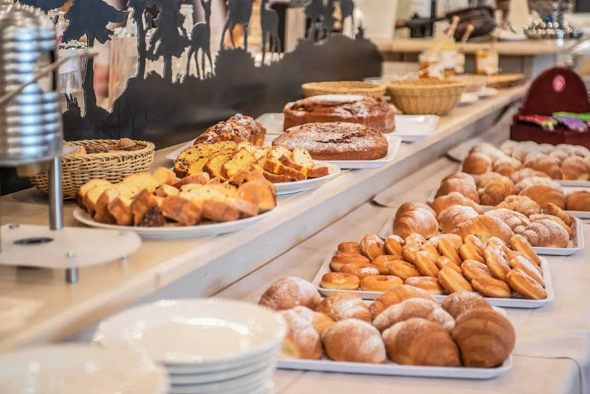 Buffet della colazione con cornetti, dolci, torte e pane fresco disposti su piatti bianchi