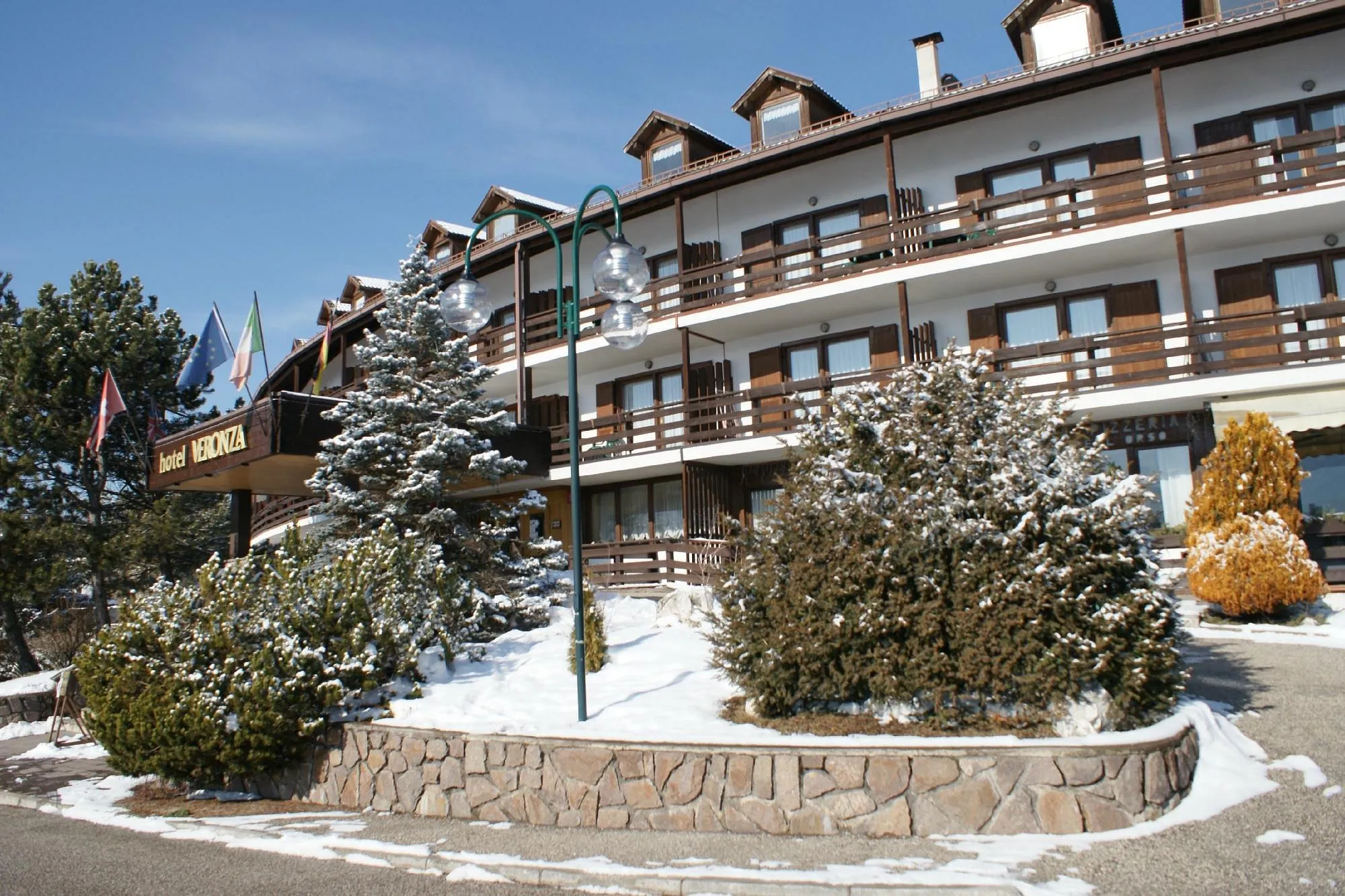 Facciata dell'Hotel Veronza in inverno con neve, abeti innevati, bandiere e insegna dell'hotel