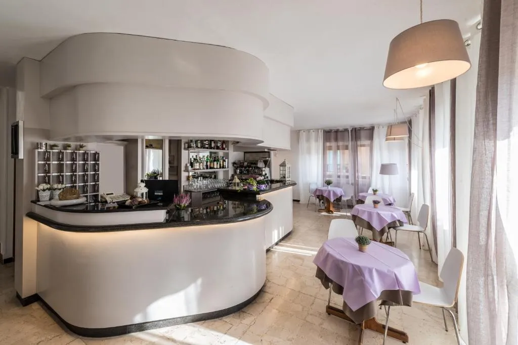 Bancone bar bianco con sala colazioni dai tavoli lilla e ampie finestre luminose