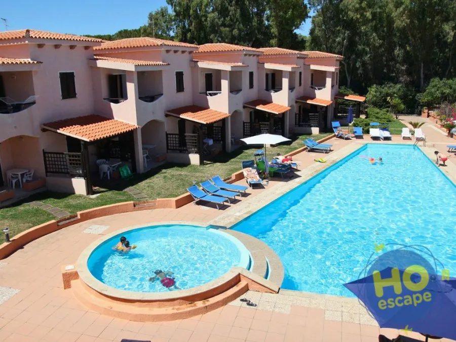 Piscina del residence con piscina per bambini, lettini blu e villette rosa immerse nel verde