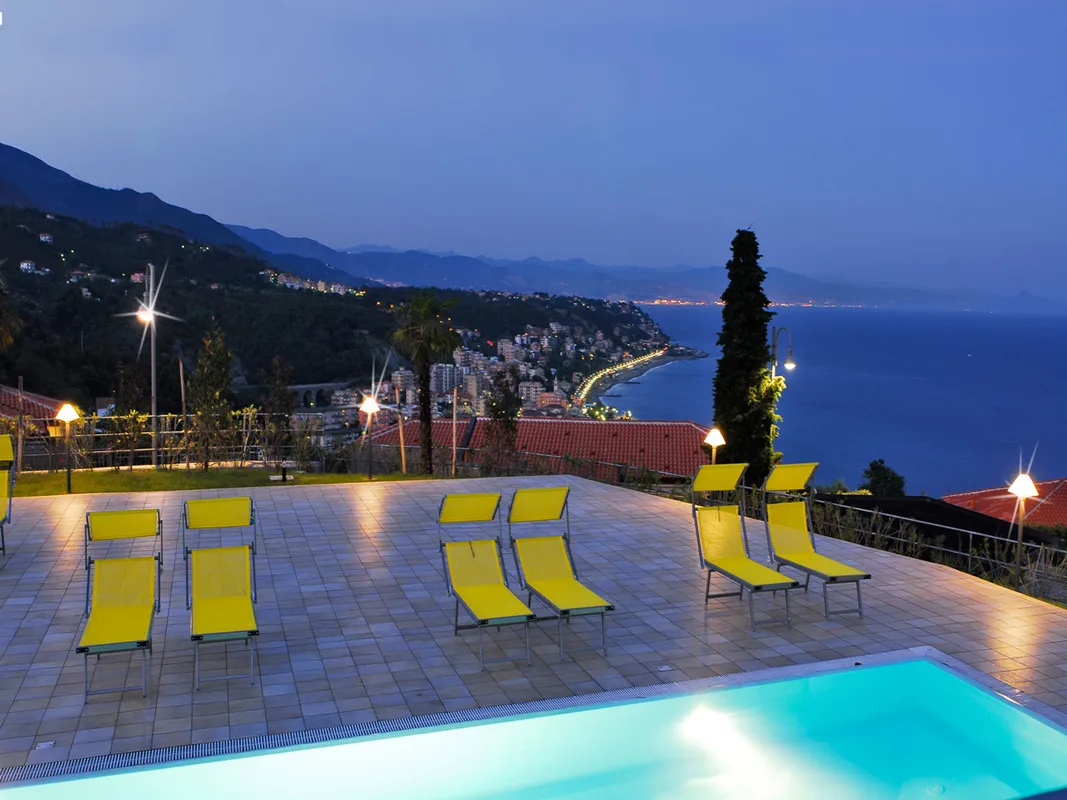 Piscina con lettini gialli al crepuscolo e vista panoramica sulle luci della costa