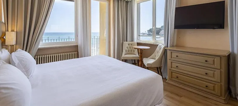 Camera con letto matrimoniale bianco, TV a parete, cassettiera e finestre con vista mare