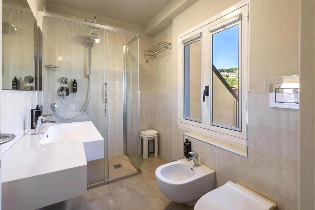 Bagno luminoso con doccia in vetro, lavabo bianco, bidet e finestra con vista sulle colline