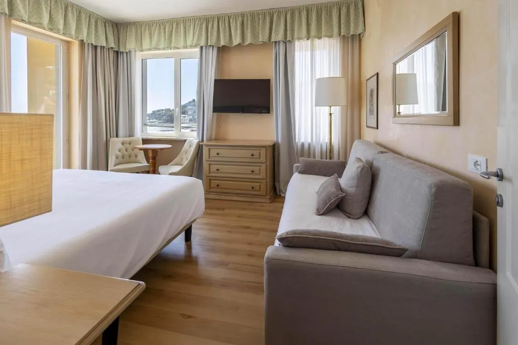 Camera con letto, divano grigio, cassettiera e poltrona accanto alla finestra con vista mare
