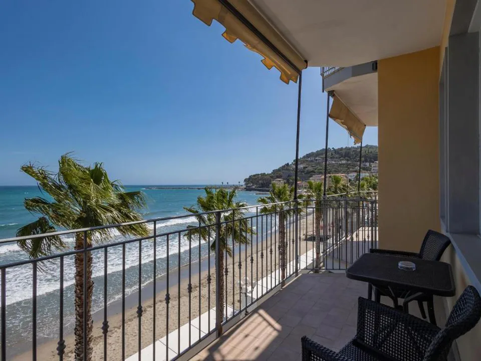 Balcone con tavolino e sedie nere affacciato su spiaggia, palme e mare azzurro con colline