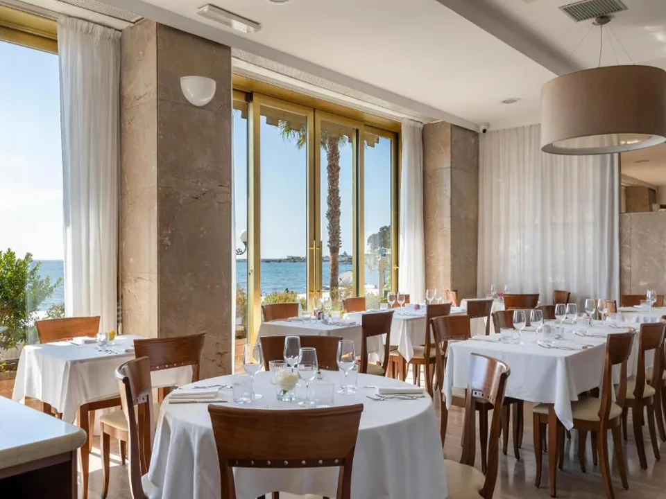 Sala ristorante luminosa con tavoli rotondi apparecchiati di bianco e vetrate con vista mare