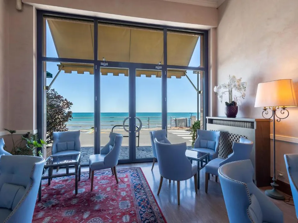 Salotto con poltrone azzurre, tappeto rosso e ampie vetrate affacciate sul mare