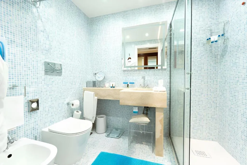 Bagno con mosaico azzurro, doccia in vetro, lavabo in pietra e tappetino blu