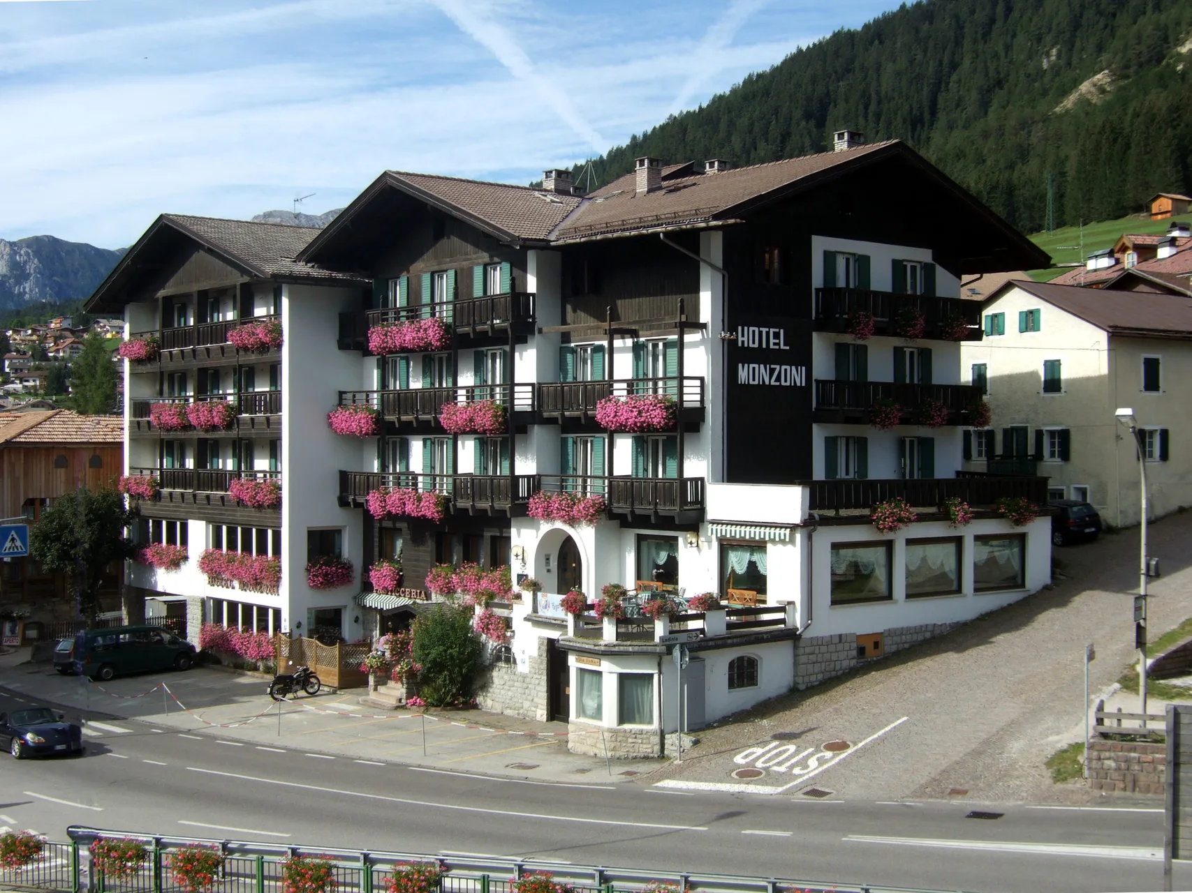 Facciata estiva dell'Hotel Monzoni con balconi fioriti di gerani rosa e montagne verdi sullo sfondo