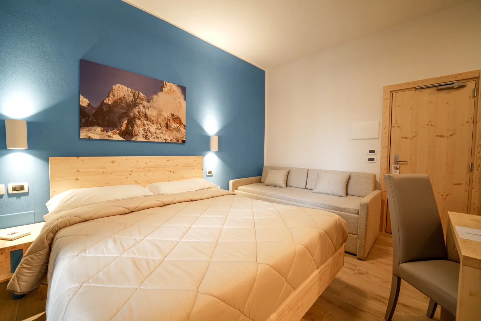 Camera matrimoniale con parete blu, letto in legno, divano e quadro delle Dolomiti