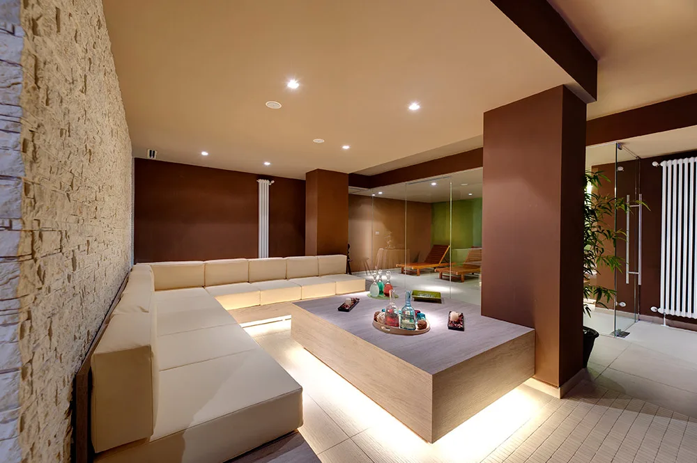 Area relax della spa con divano in pelle chiara, parete in pietra e tavolo con prodotti benessere