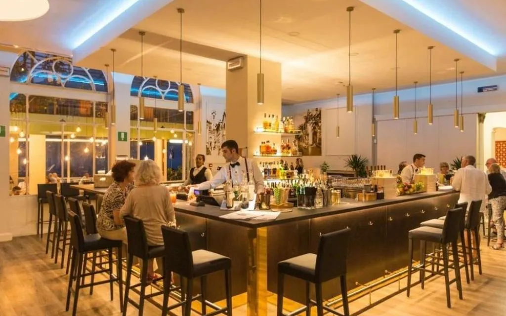 Bar dell'hotel con bancone illuminato, barman e clienti seduti su sgabelli
