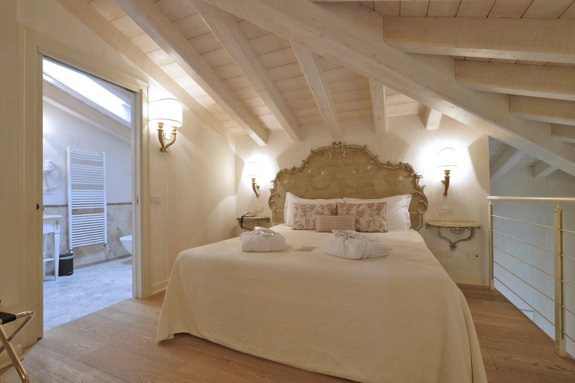 Camera mansardata con travi in legno chiaro, letto matrimoniale con testiera decorata e bagno privato