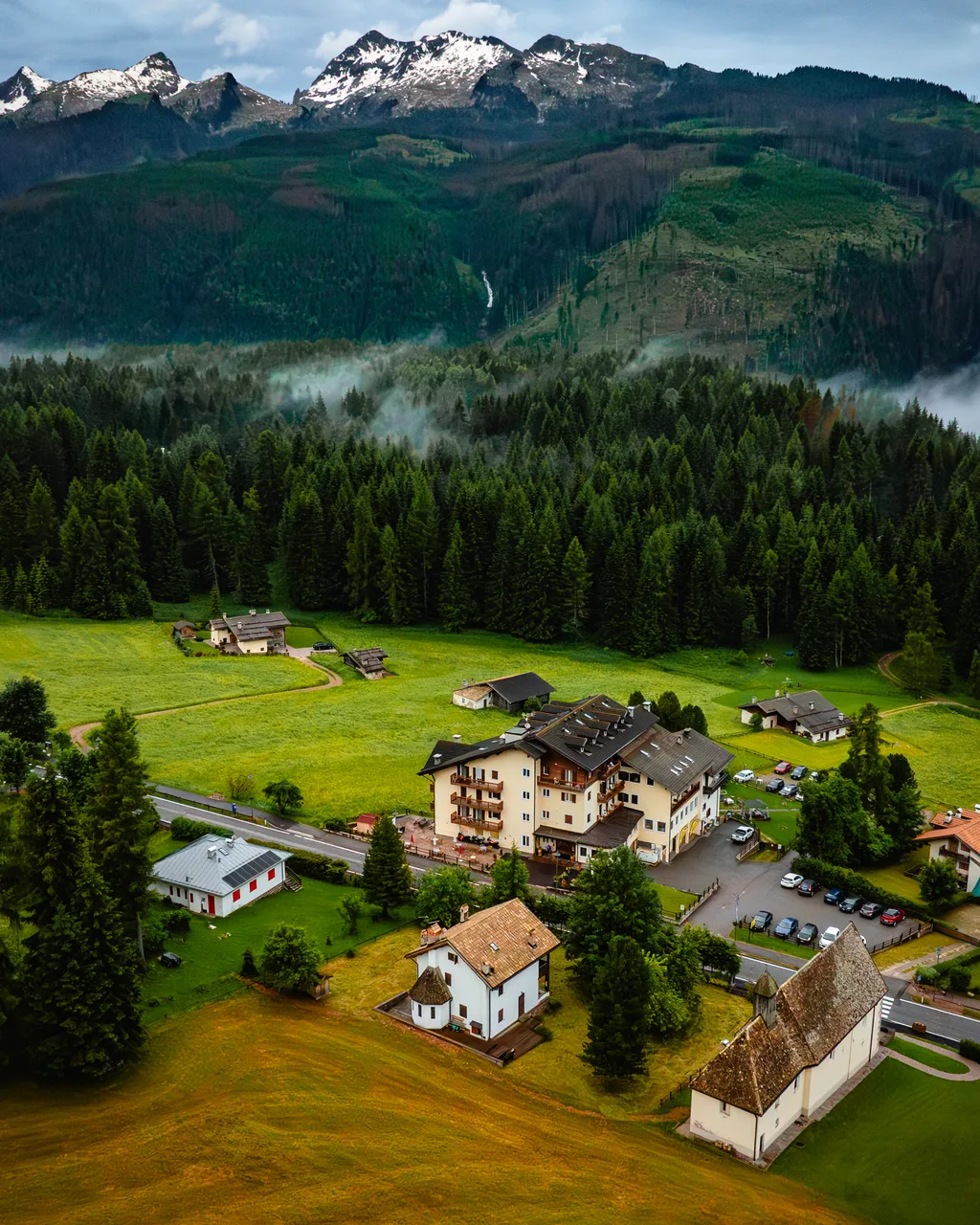 Veduta aerea dell'hotel tra prati e foreste con montagne innevate e nebbia sullo sfondo