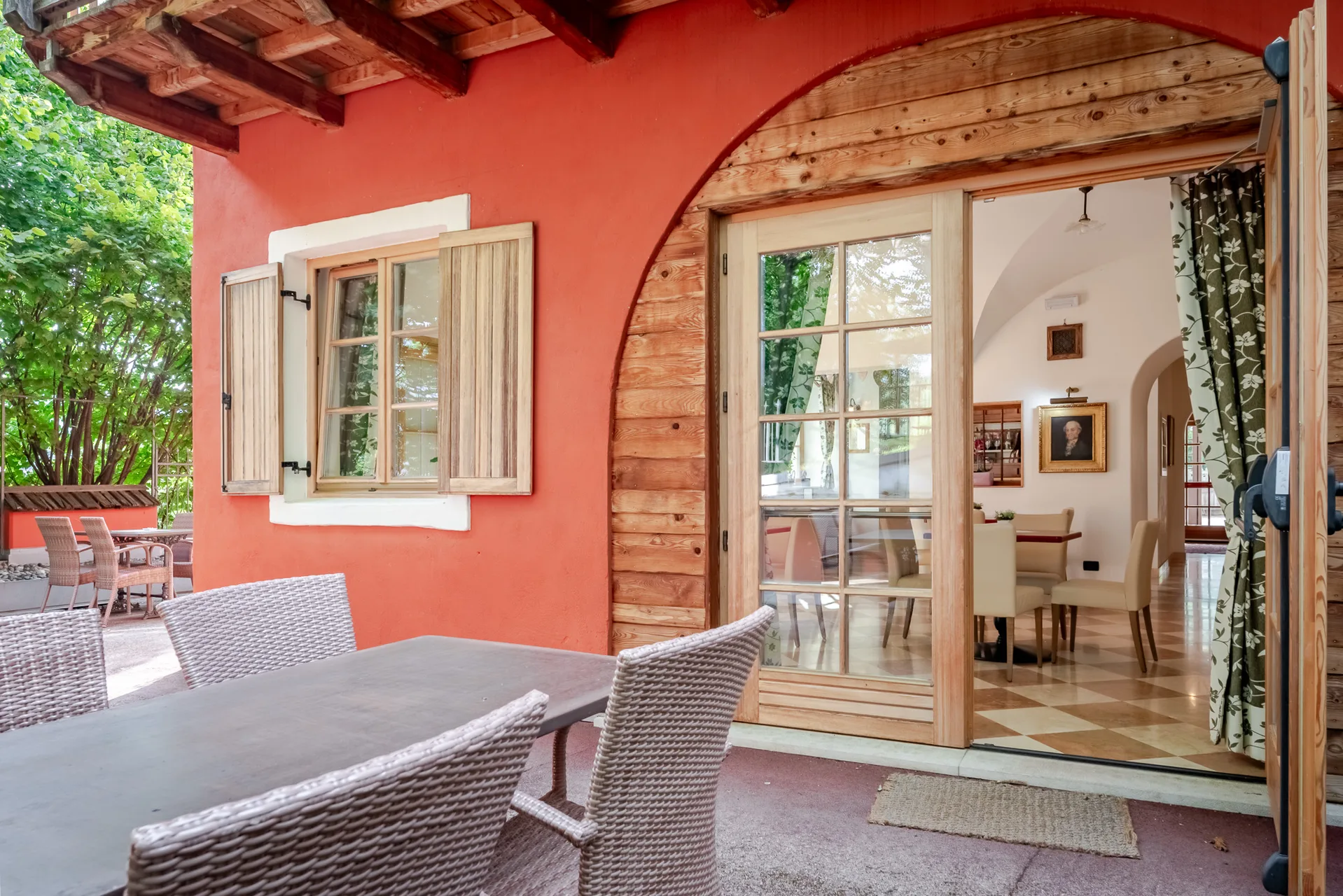 Portico esterno con parete rossa, tavolo e sedie in vimini affacciato sulla sala da pranzo