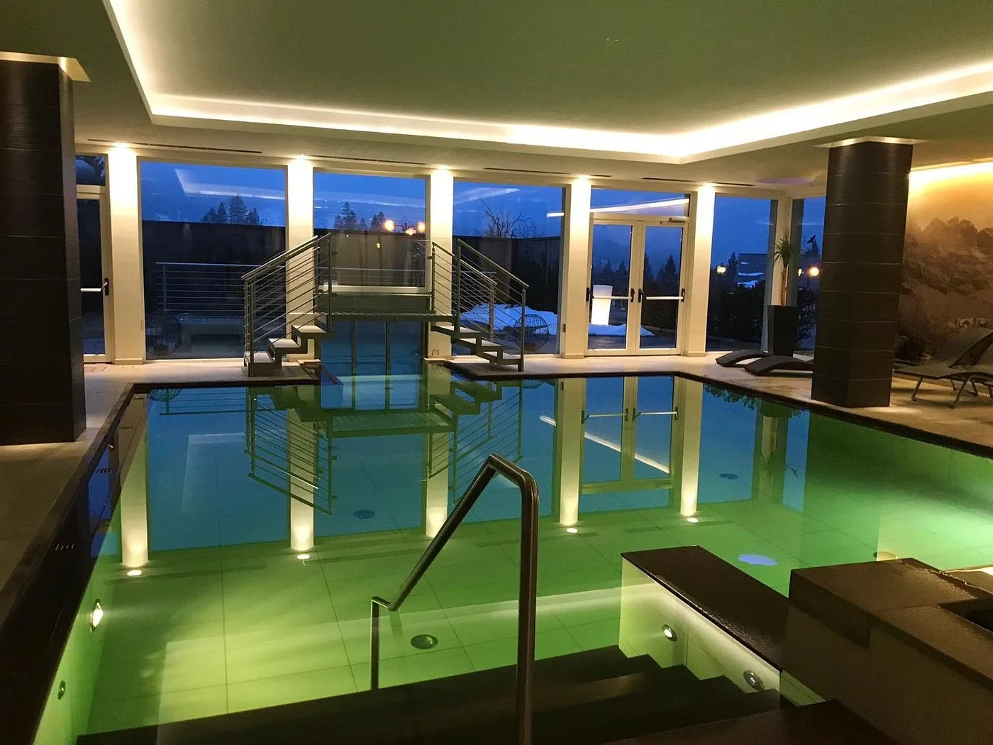 Piscina interna con illuminazione verde e grandi vetrate al crepuscolo