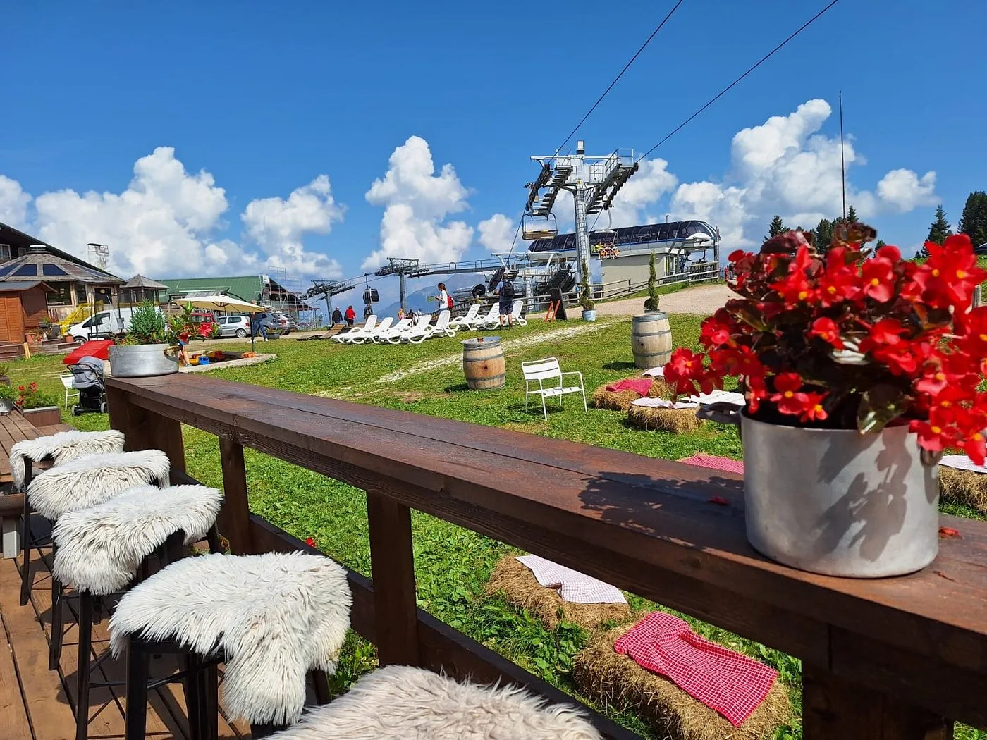 Bar esterno con sgabelli in pelliccia, fiori rossi e vista sulla seggiovia estiva