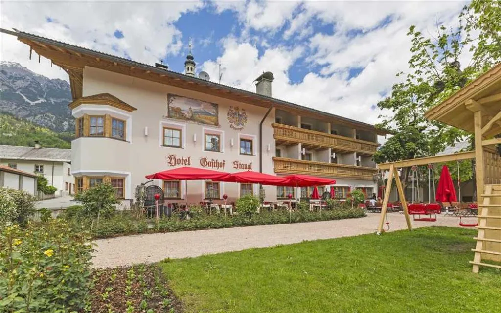 Esterno dell'hotel in estate con giardino, terrazza con ombrelloni rossi e area giochi per bambini