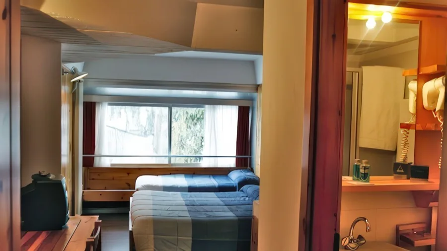Camera d'albergo con due letti singoli, grande finestra con tende, televisore su mobile e bagno con lavabo e specchio.