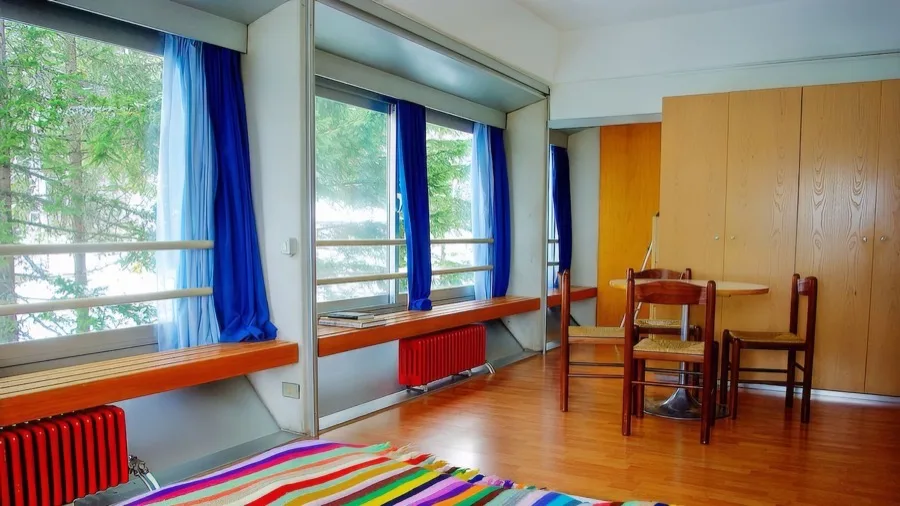 Interno luminoso con grandi finestre, tende blu, tavolo rotondo con quattro sedie e parquet con tappeto colorato.