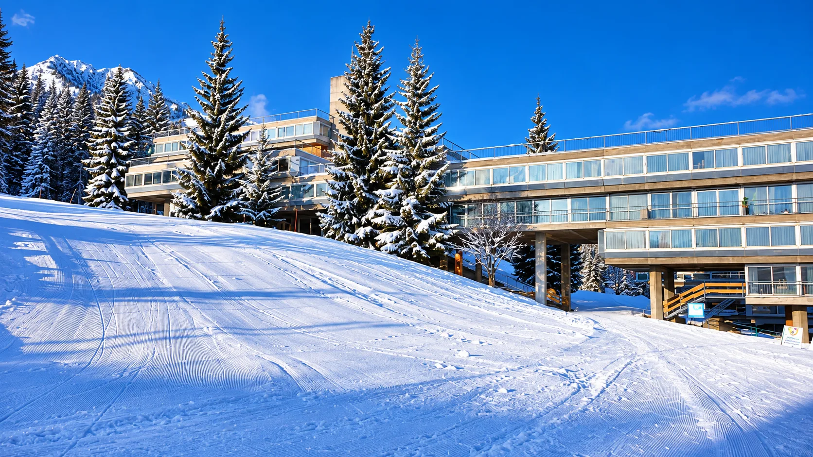 Hotel a bordo pista con pini innevati e montagna sullo sfondo in giornata di sole