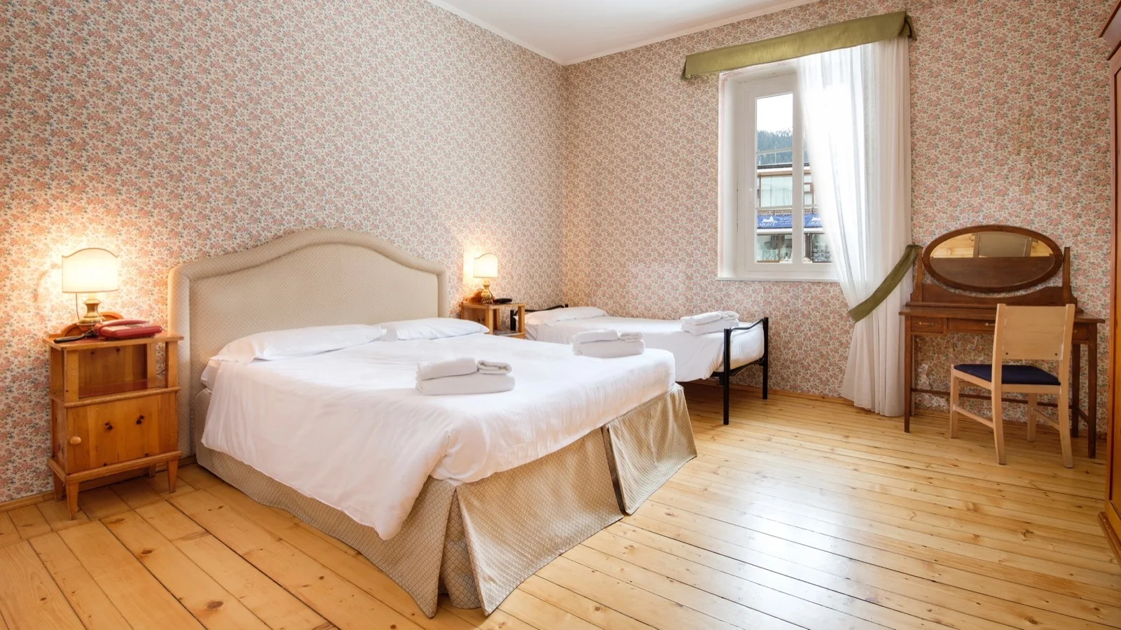Camera da letto con due letti, mobili in legno, pareti con carta da parati floreale e pavimento in legno chiaro.