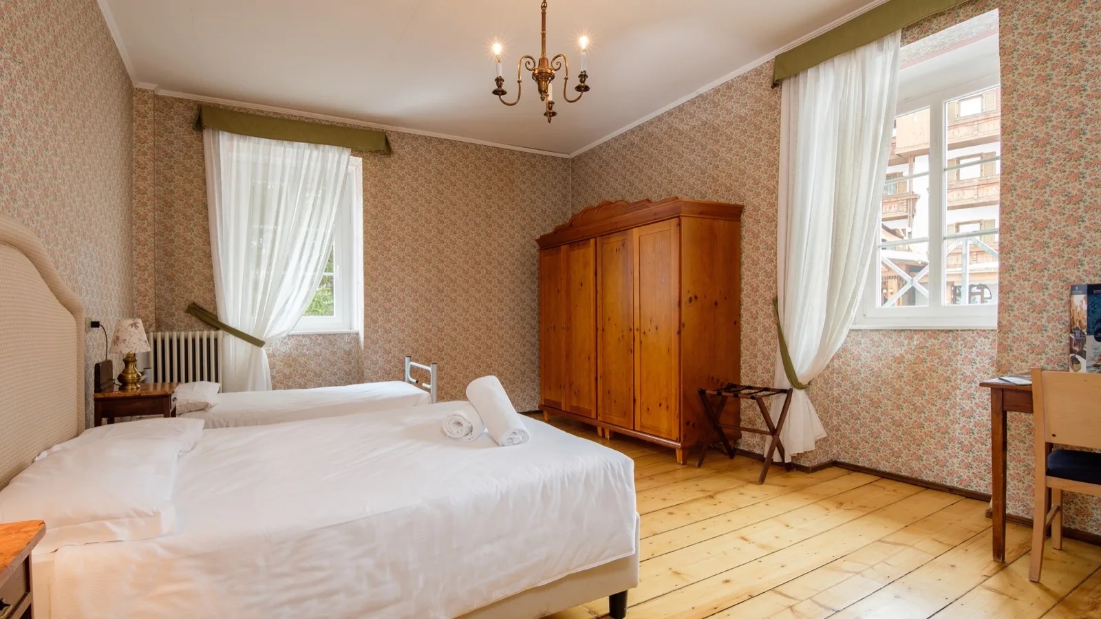 Camera matrimoniale con letto in primo piano, armadio in legno, pavimento in parquet e finestre con tende bianche.