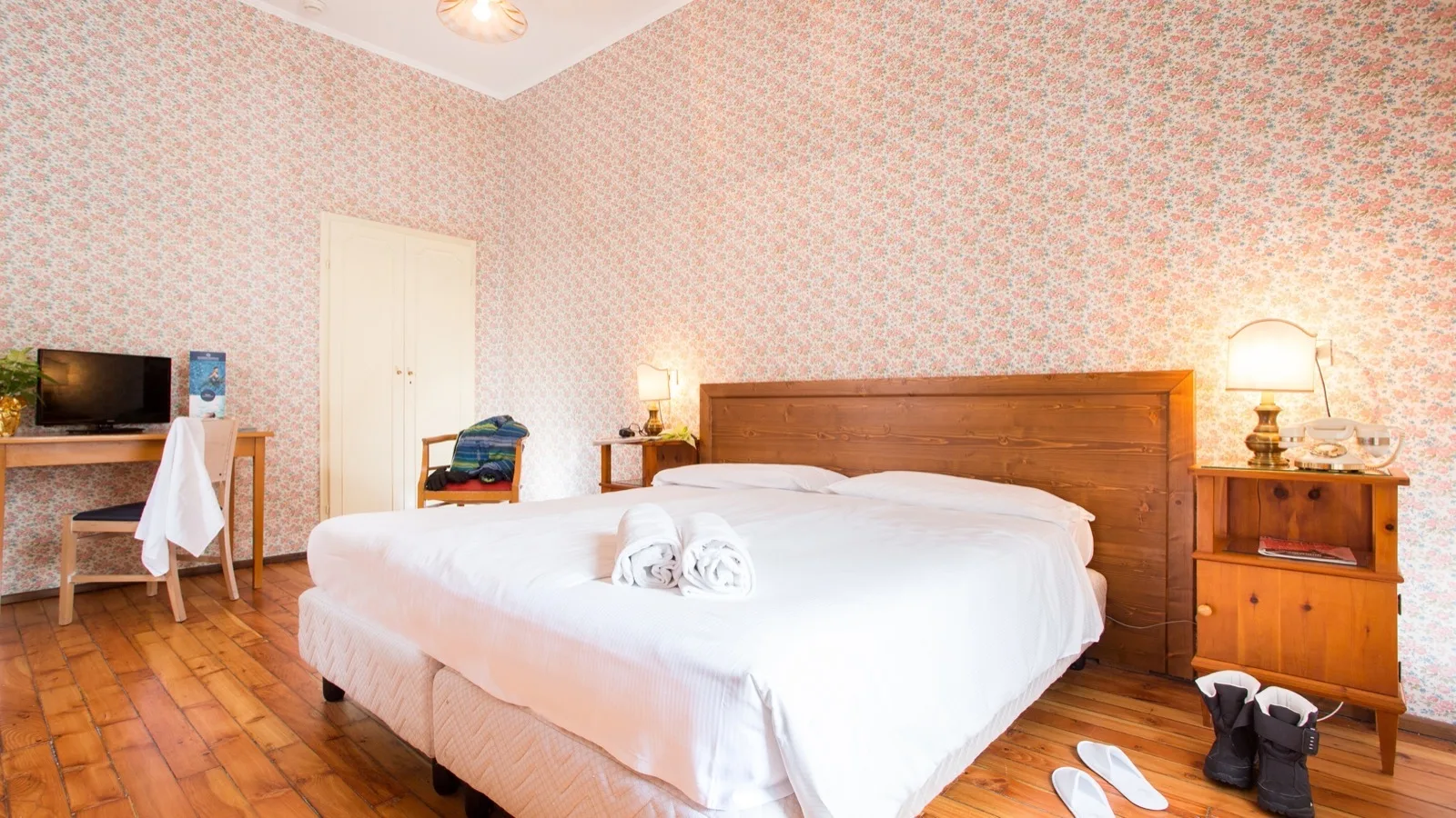 Camera da letto con letto matrimoniale, pareti a carta da parati floreale, pavimento in legno, scrivania, e stivali da neve accanto al comodino.