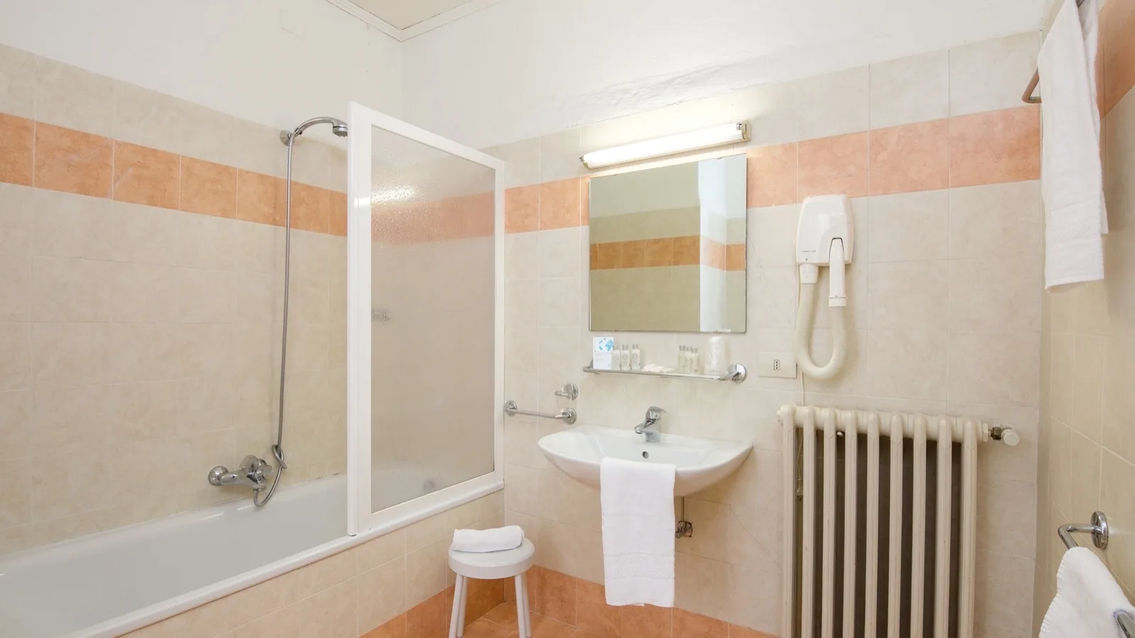 Bagno con vasca e doccia, lavabo con specchio, asciugacapelli e scaldasalviette, piastrelle beige e arancioni.