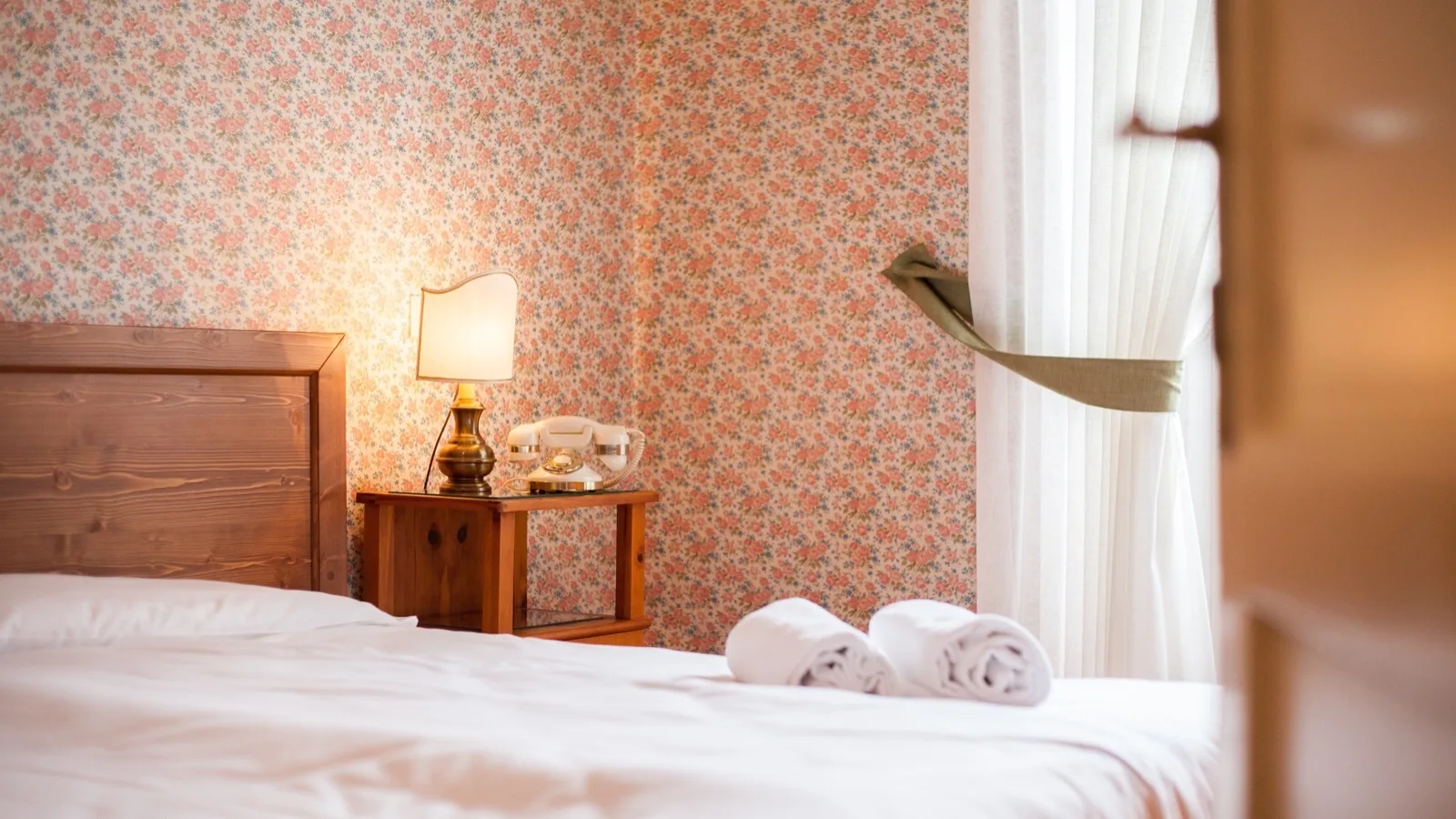 Camera da letto con letto in legno, pareti con carta da parati floreale, lampada accesa e telefono vintage su comodino.