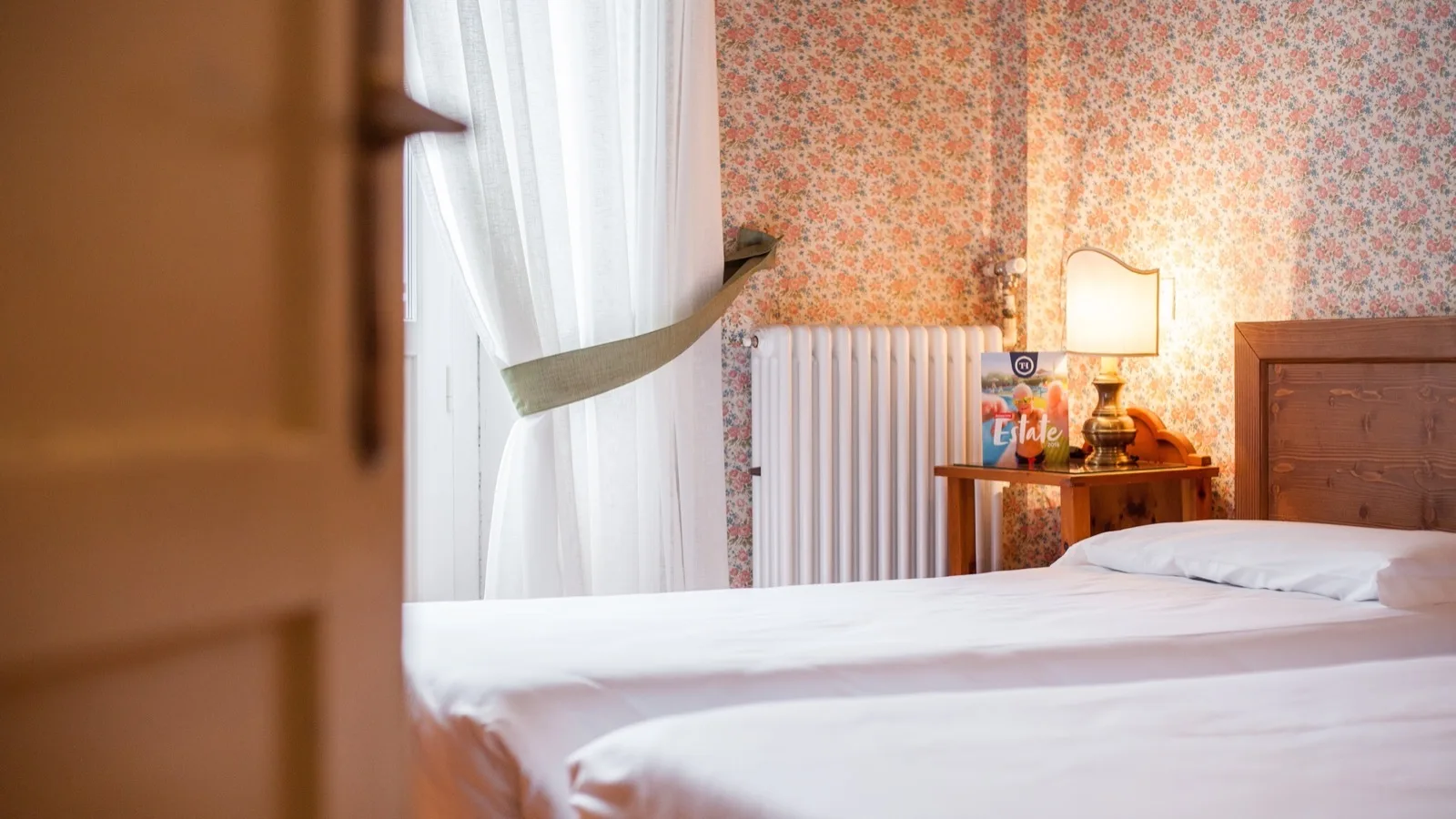Camera da letto con letto singolo, carta da parati floreale, una tenda bianca e una lampada su comodino in legno.