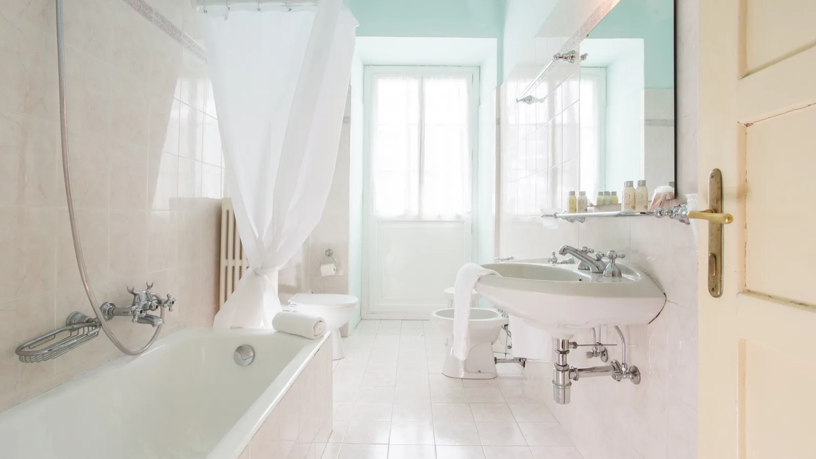 Bagno luminoso con vasca, doccia con tenda bianca, lavabo, bidet e wc, pavimento piastrellato chiaro e finestra sullo sfondo.