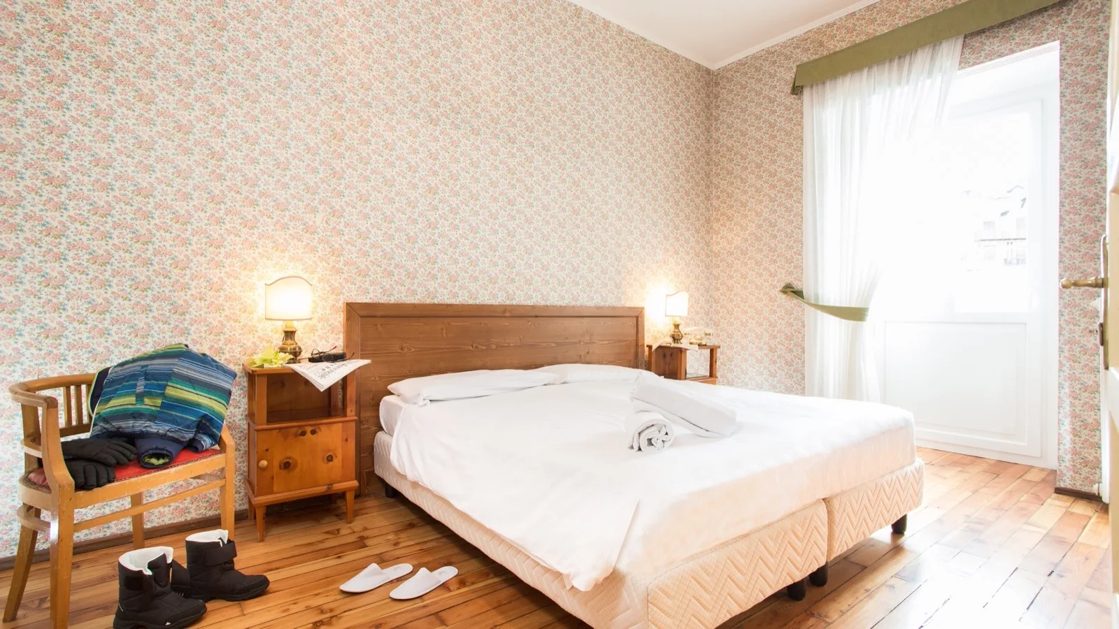Camera da letto luminosa con letto matrimoniale, carta da parati floreale, pavimento in legno e finestra con tenda bianca.