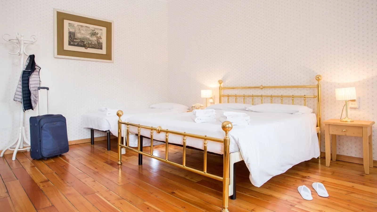 Camera da letto con letto matrimoniale in ottone, comodino con lampada, appendiabiti con giacca e valigia blu.