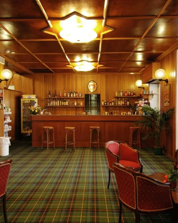 Interno di bar con pareti e soffitto in legno, bancone con sgabelli, poltrone rosse e moquette a quadri verdi.