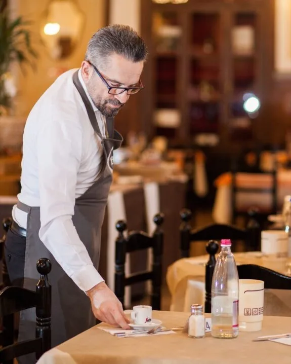 Cameriere con occhiali e grembiule che sistema una tazzina di caffè su un tavolo di ristorante.