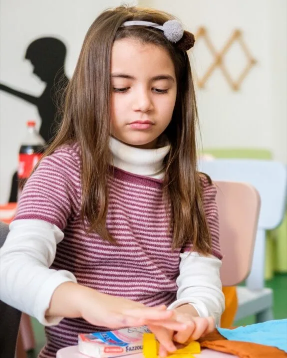 Bambina con fascia per capelli mentre gioca con pastelli gialli su un tavolo colorato in ambiente interno.
