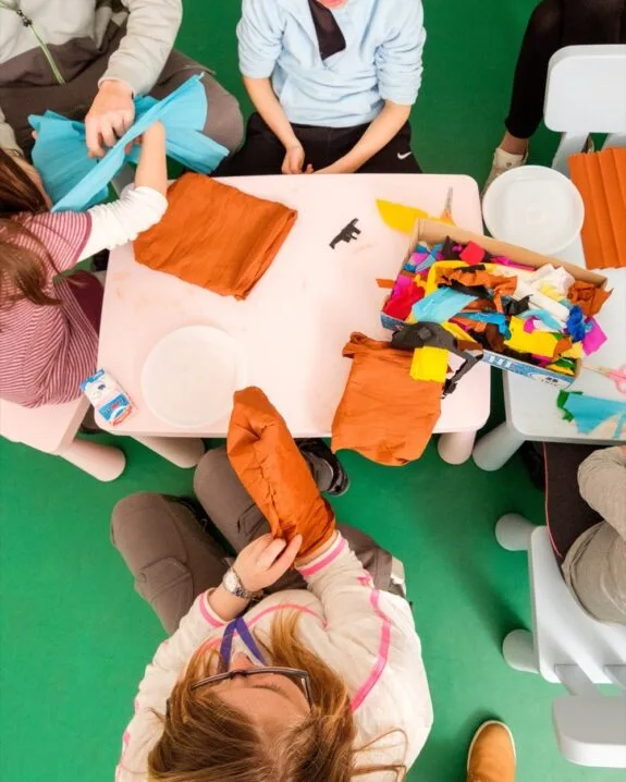 Bambini e un adulto seduti intorno a un tavolo colorato con carta velina e materiali per attività creative.