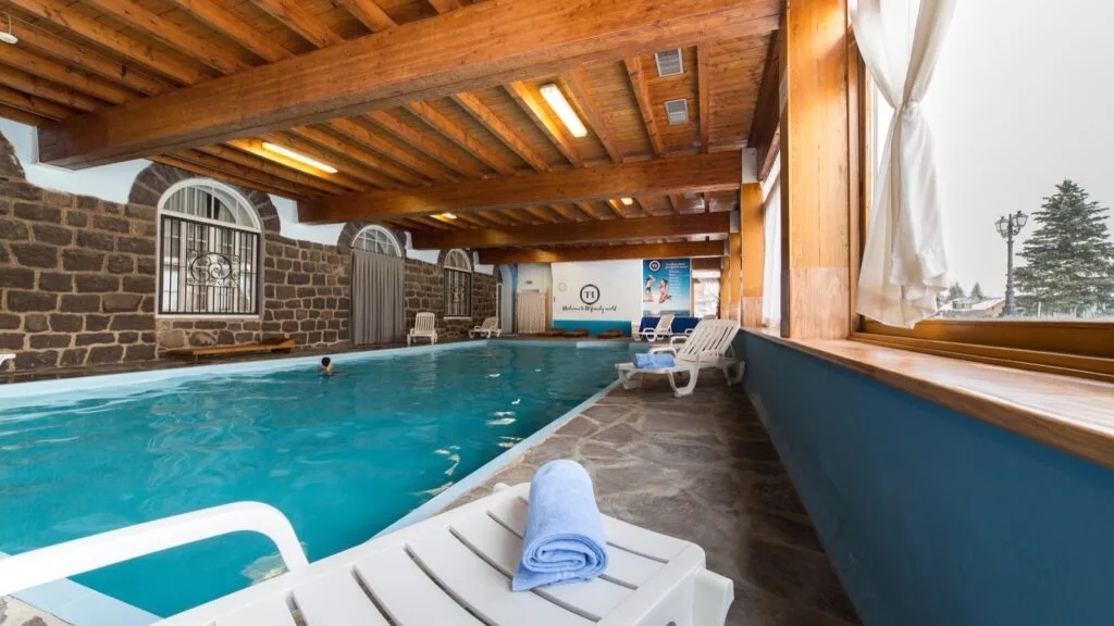 Piscina coperta con pareti in pietra, soffitto in legno, sedie a sdraio bianche e finestra con tenda bianca.