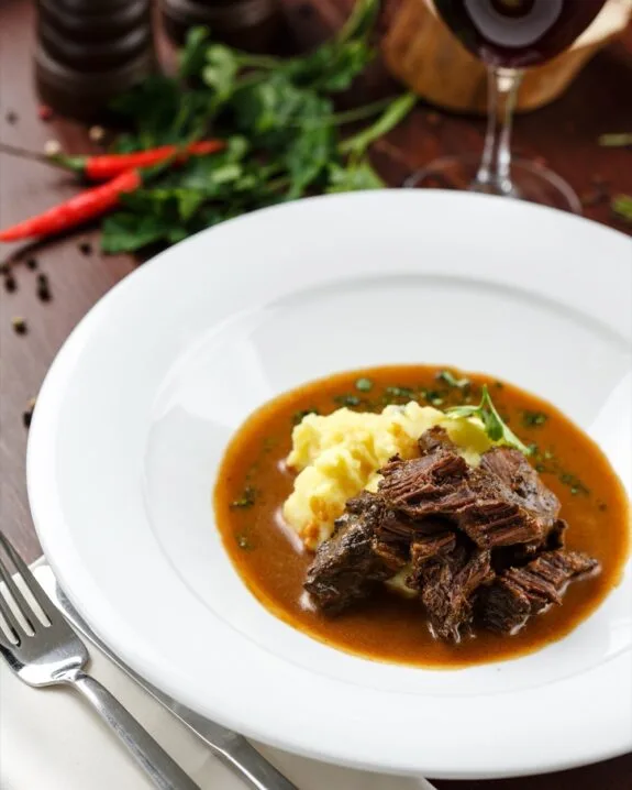 Piatto bianco con spezzatino di carne in salsa marrone e purè di patate guarnito con prezzemolo.
