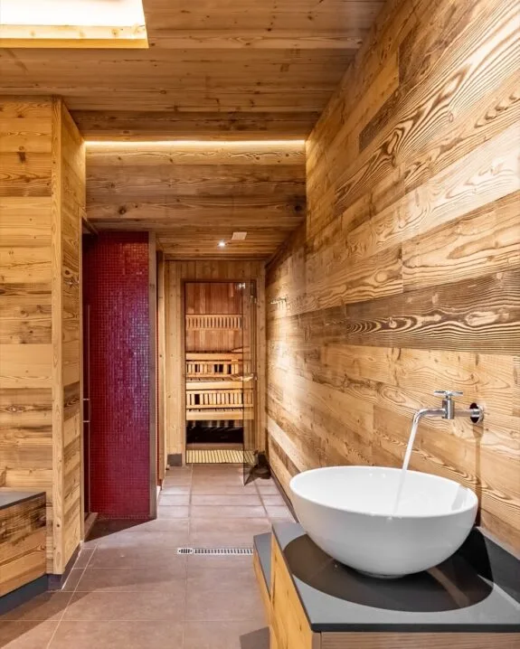 Interno di sauna in legno con lavabo bianco e rubinetto, pavimento in piastrelle e parete con piastrelle rosse.