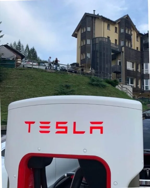 Colonnina di ricarica Tesla bianca con logo rosso in primo piano, edificio e persone sullo sfondo erboso.