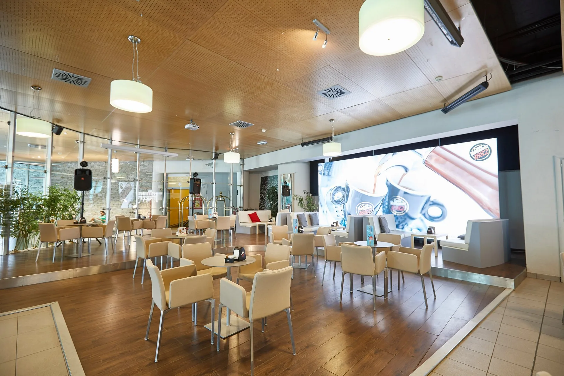 Sala lounge con tavolini, sedie bianche e grande schermo pubblicitario