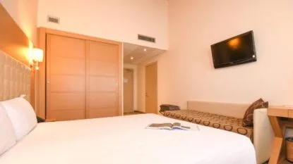 Camera d'albergo luminosa con letto matrimoniale, divano, televisore a muro e armadio con porte scorrevoli in legno.