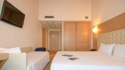 Camera d'hotel con letto matrimoniale, divano letto singolo, armadio in legno, TV al muro e illuminazione calda.